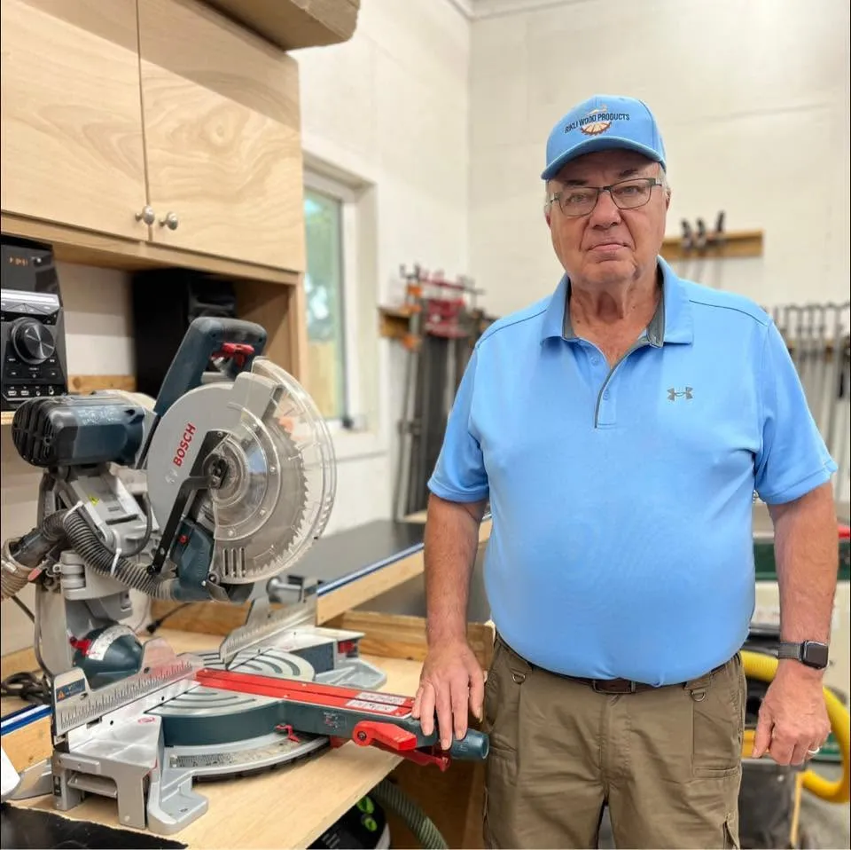 Owner of Rikli Wood Products standing in workshop next to woodworking saw and equipment, wearing branded hat and shirt.